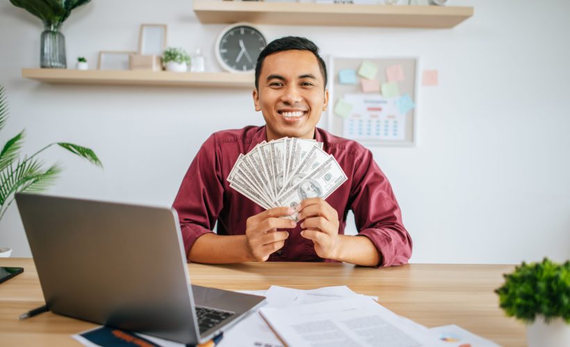 a man working in an office and holding money, with a laptop documents on the desk.